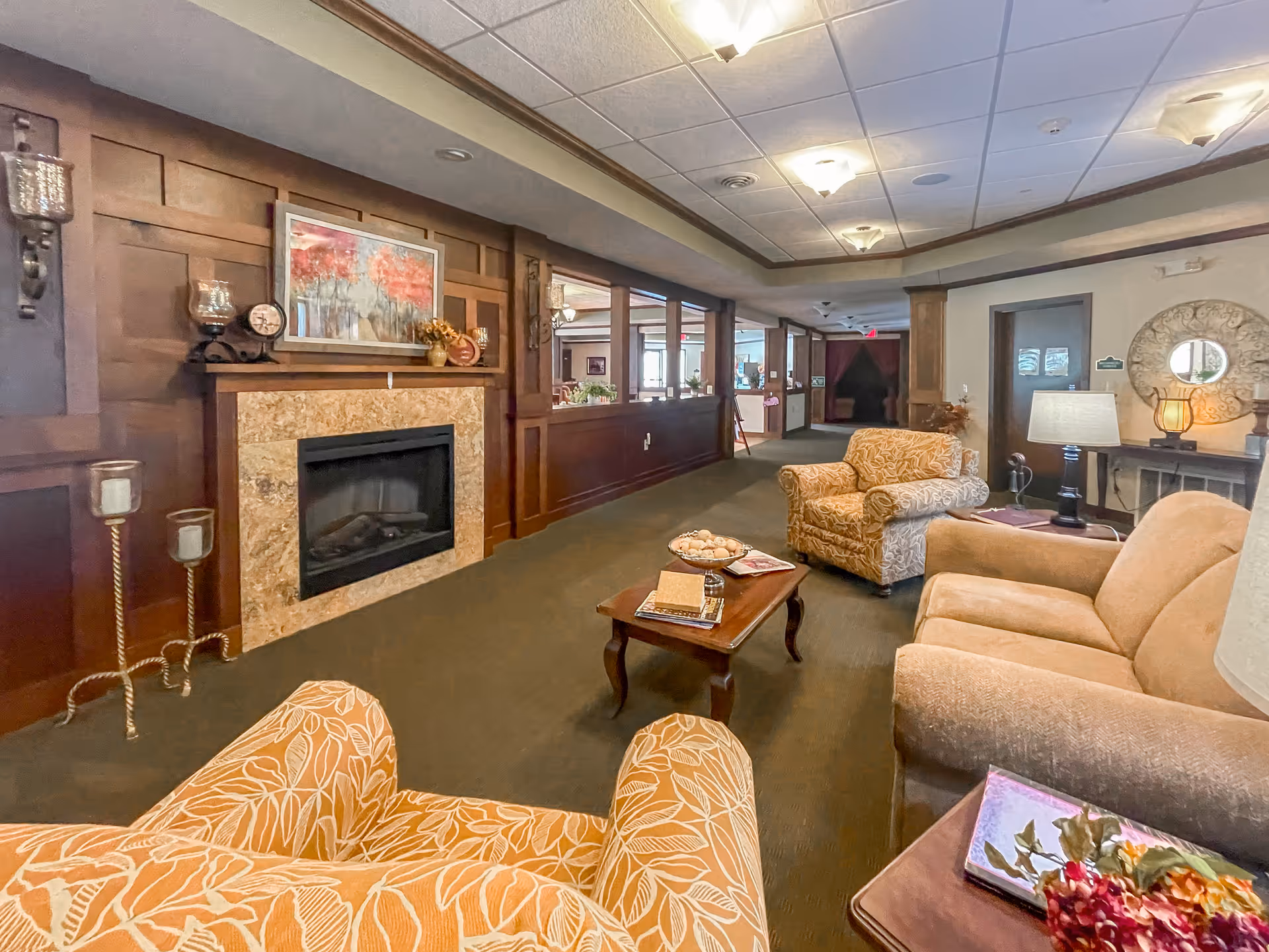 A cozy assisted living facility common area with comfortable upholstered armchairs and a sofa arranged around a wooden coffee table. The room features a decorative fireplace with a painting above it, candle holders, and warm wood paneling on the walls. Soft lighting from ceiling fixtures and table lamps creates a welcoming atmosphere.