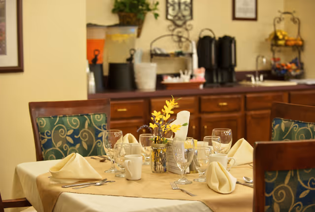A dining table set for four with beige napkins, glassware, white coffee mugs, and silverware. A small vase with yellow flowers is in the center of the table. In the background, there is a counter with beverage dispensers and a decorative wall hanging.