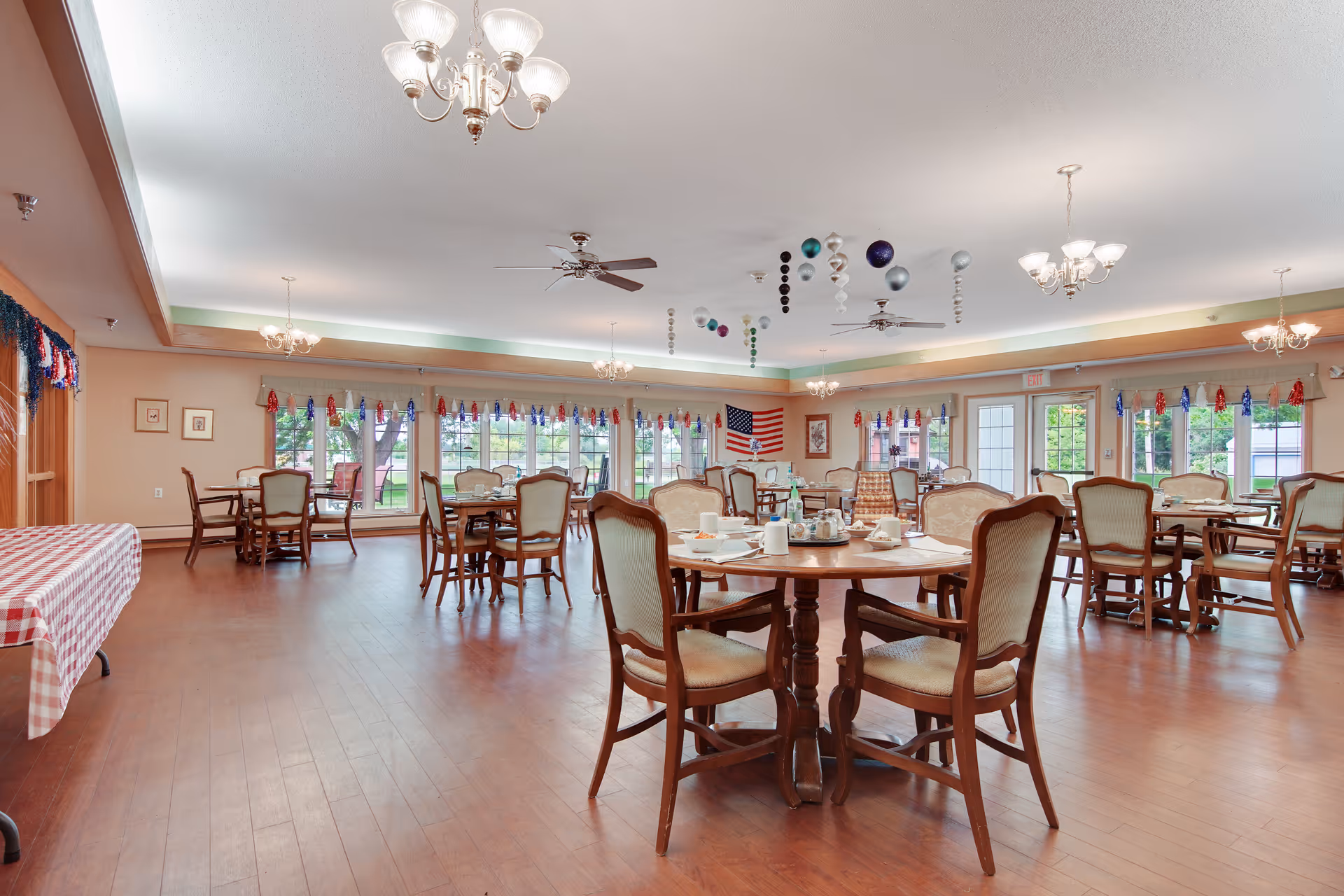 A spacious dining room with multiple round wooden tables and cushioned chairs arranged neatly. The room has large windows with red, white, and blue decorations hanging above them, and an American flag displayed on the far wall. The ceiling features chandeliers and ceiling fans, and the floor is wooden. A table with a red and white checkered tablecloth is visible on the left side.