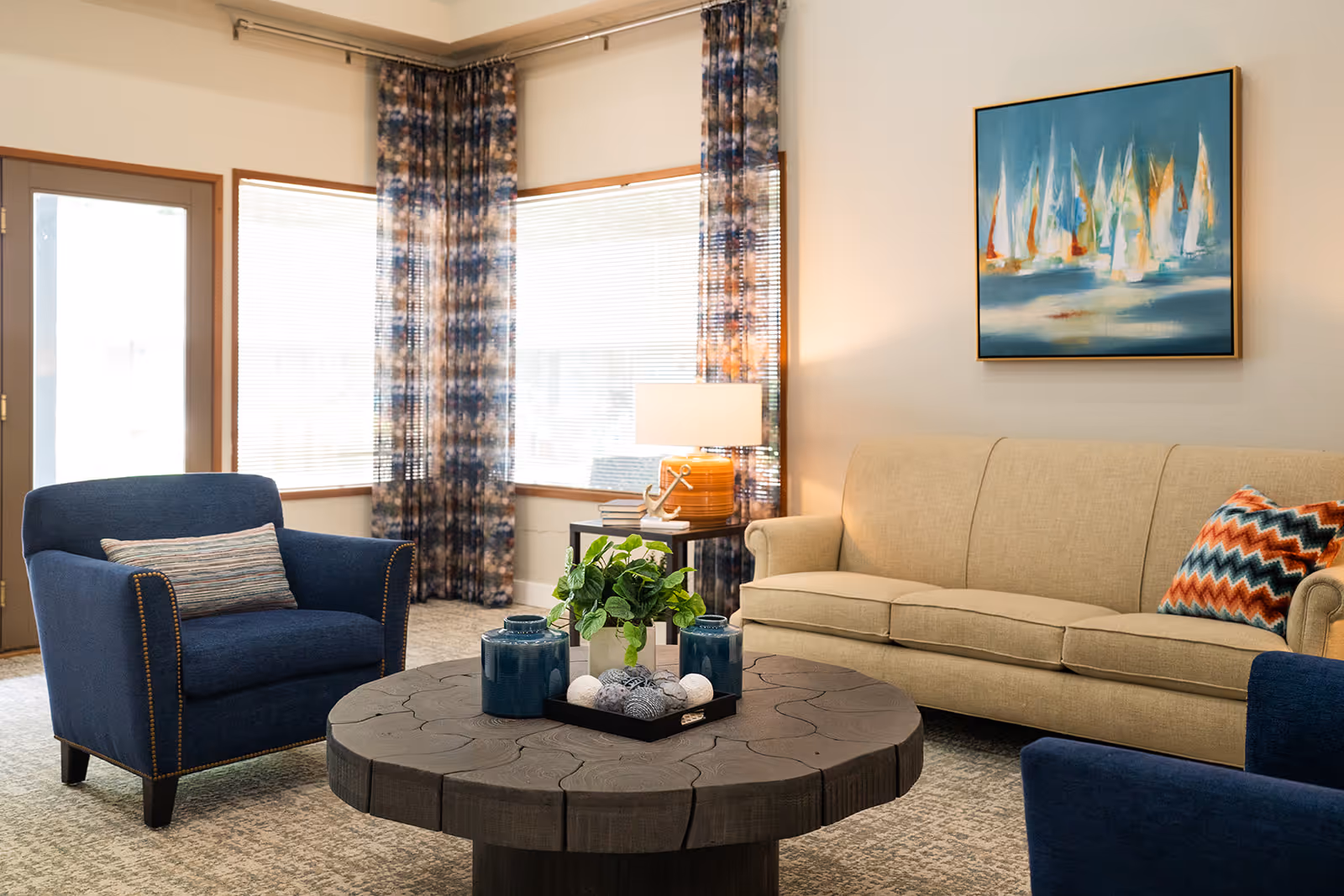 A cozy living room with a beige sofa adorned with a colorful zigzag patterned pillow, two blue armchairs with a striped pillow on one, a round wooden coffee table with decorative items including blue vases and a plant, a side table with a lamp and books, large windows with blinds and patterned curtains, and a painting of sailboats on the wall.
