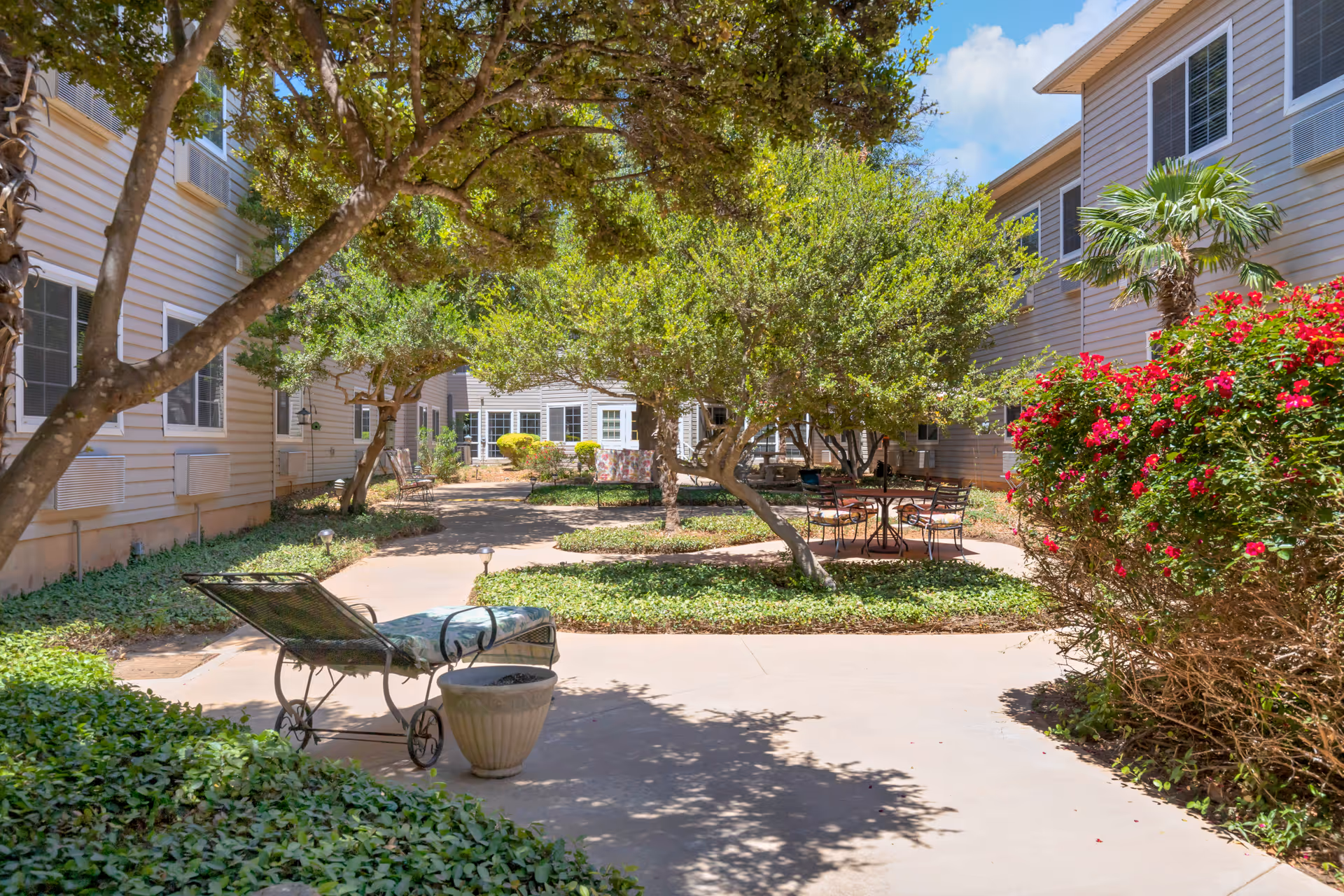 Sunlit courtyard with trees, patio chairs and a lounge chair between two wings of a senior living facility.