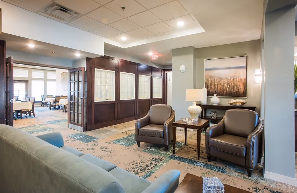 A cozy common area in a senior living facility featuring two brown leather armchairs and a green sofa arranged around a wooden side table with a lamp. The room has patterned carpet, soft lighting, and a large painting on the wall. In the background, there is a dining area with tables and chairs visible through open double doors.
