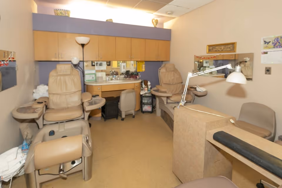 A small room with two beige pedicure chairs equipped with foot baths, a sink with cabinets above, a floor lamp, a desk lamp, and a chair. The walls are light-colored with a purple accent behind the sink area, and there are mirrors and a calendar on the walls.