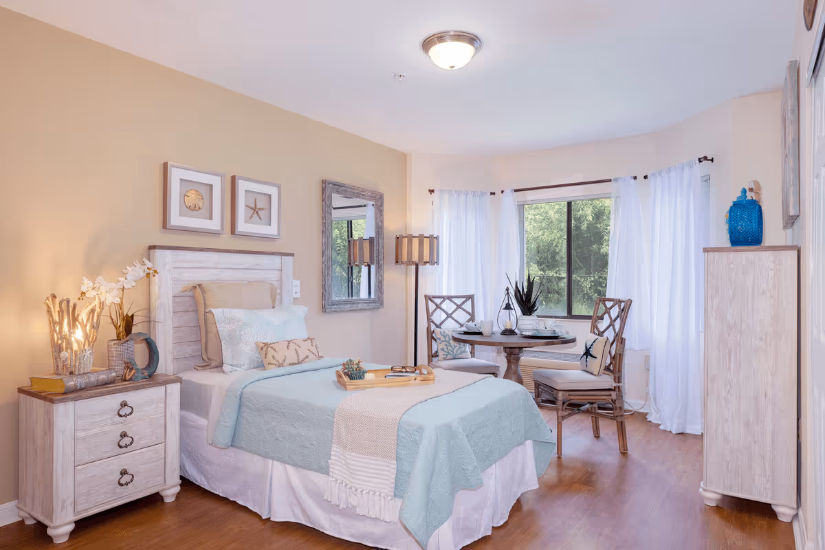 A cozy bedroom with a single bed dressed in light blue and beige bedding. Next to the bed is a wooden nightstand with decorative items including a lamp, books, and a small plant. On the wall above the bed are two framed pictures and a mirror. In the corner near a window with white curtains is a small round table set for two with chairs, decorated with cushions and a plant. The room has wooden flooring and soft lighting.