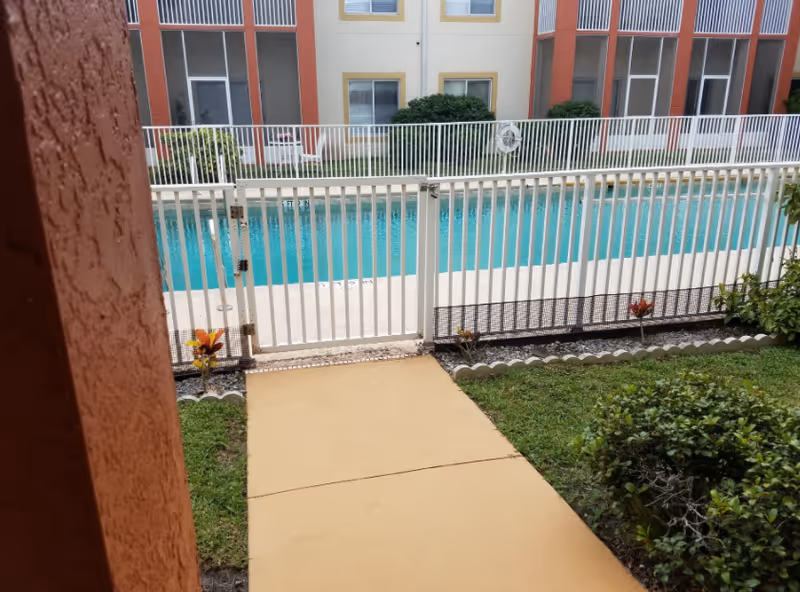 View of a swimming pool enclosed by a white metal fence with a gate. The pool is surrounded by a concrete deck and is located in the courtyard of a multi-story residential building with screened balconies and windows. There are small landscaped areas with grass, bushes, and flowers near the walkway leading to the pool gate.