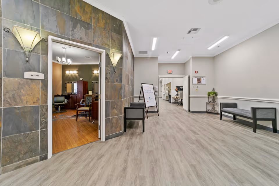 Interior hallway of a senior living facility with light wood flooring and gray walls. On the left, there is an open doorway leading to a salon with salon chairs and mirrors. The hallway has benches along the right wall and a signboard in the middle. The ceiling has recessed lighting and an exit sign is visible at the far end.