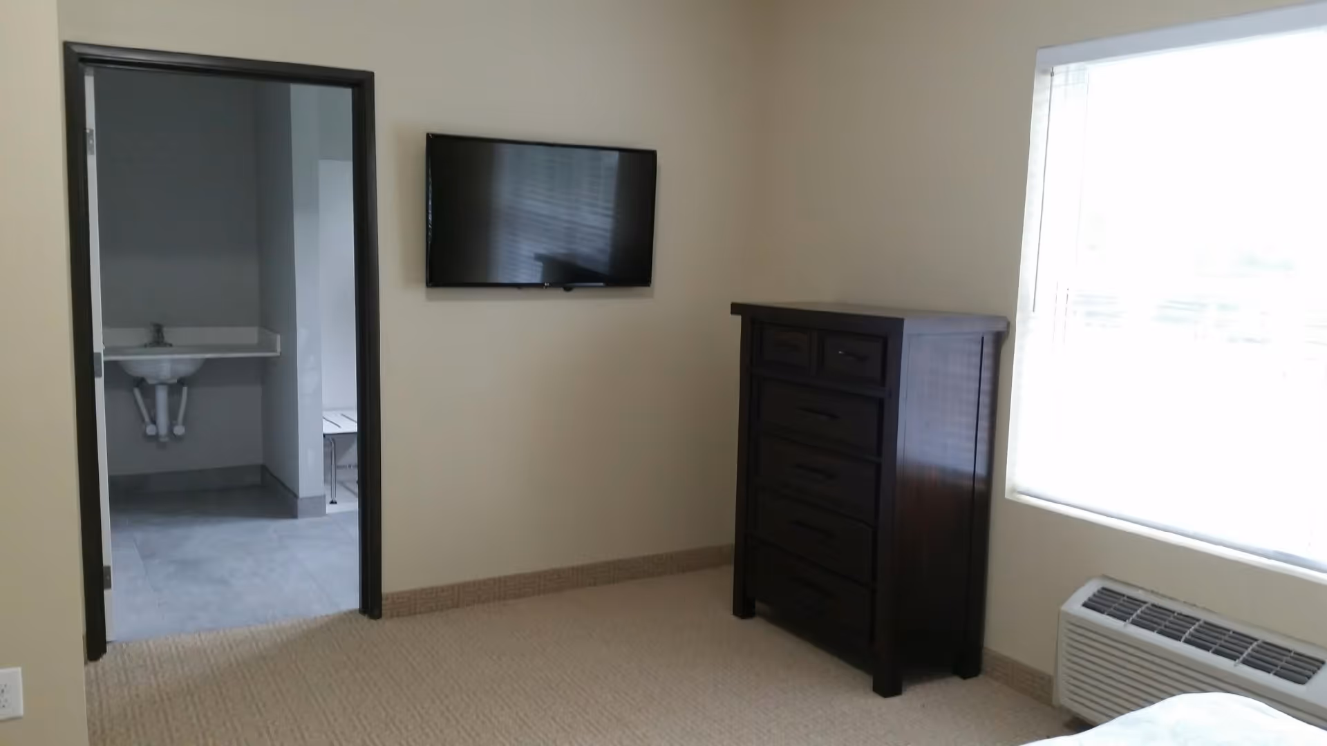 A simple room with beige walls and carpet, featuring a dark wooden dresser, a wall-mounted flat screen TV, a large window with blinds, and a view into an adjacent bathroom with a sink and tiled floor.