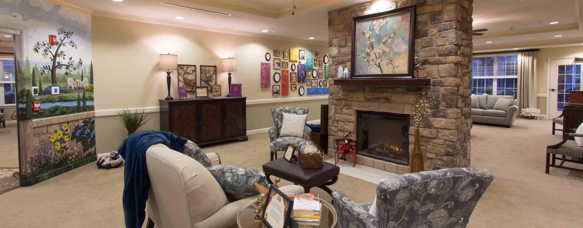 A cozy living room area in a senior living facility featuring a stone fireplace with a painting above it, surrounded by comfortable upholstered chairs and a small ottoman. The room has warm lighting from table lamps on a wooden sideboard against the wall, decorated with various framed pictures and artwork. A colorful mural with nature scenes is visible on a partial wall to the left, and large windows with curtains allow natural light into the space.