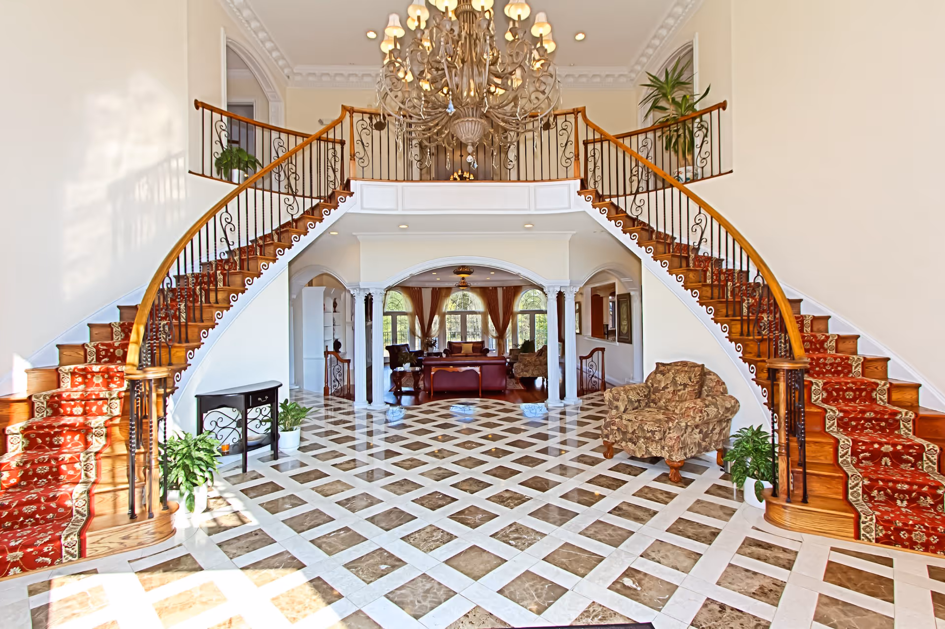 A grand interior space featuring a double curved staircase with red carpet runners and ornate black railings. The floor is tiled with a checkered pattern of brown and white marble. A large chandelier hangs from the ceiling above the staircase. In the background, there is a sitting area with sofas, armchairs, and large windows with curtains allowing natural light to fill the room. Several potted plants are placed around the space.