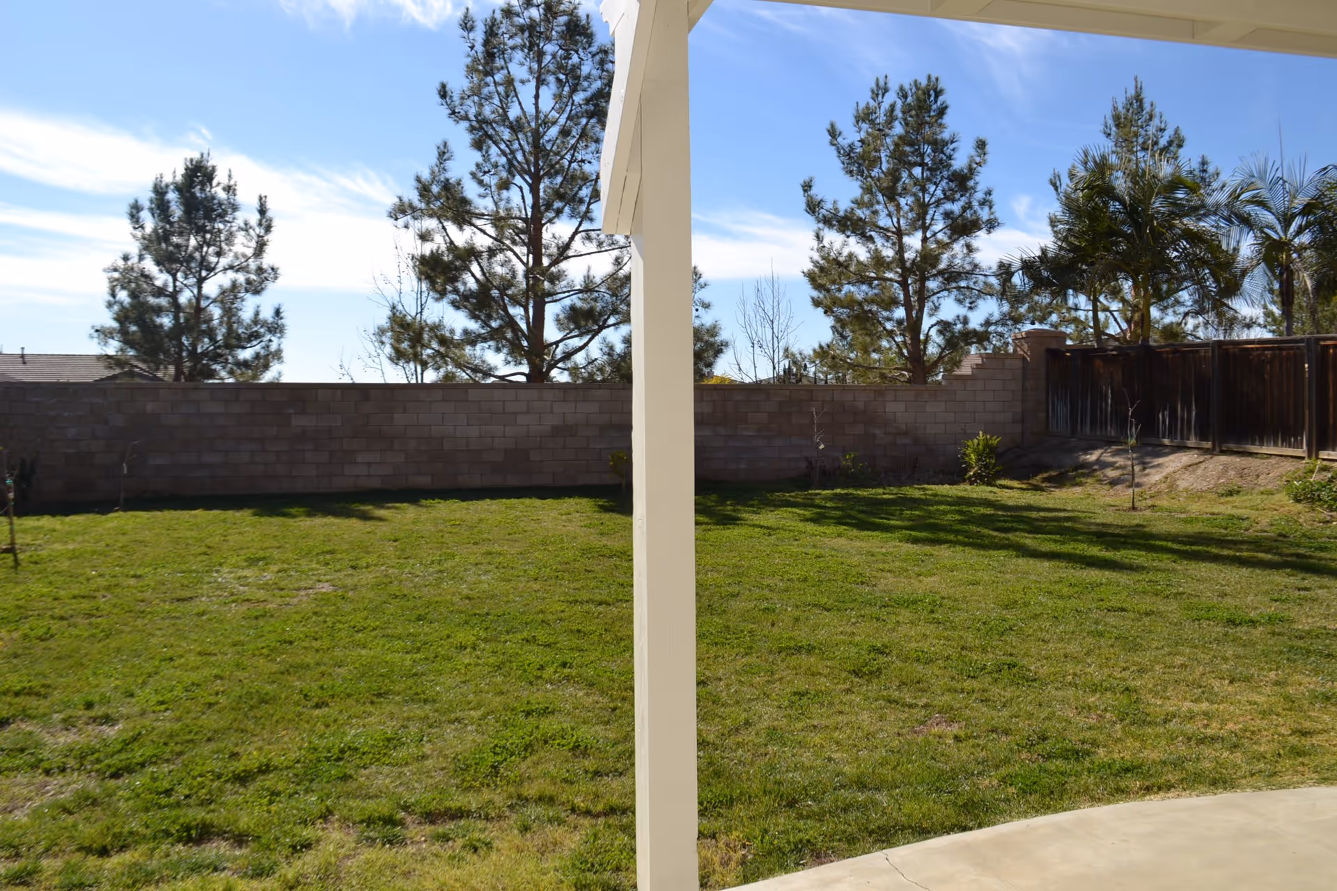 View of a backyard with green grass, a concrete patio, a white support post in the center, a brick wall fence, wooden fence, and several trees under a blue sky with some clouds.