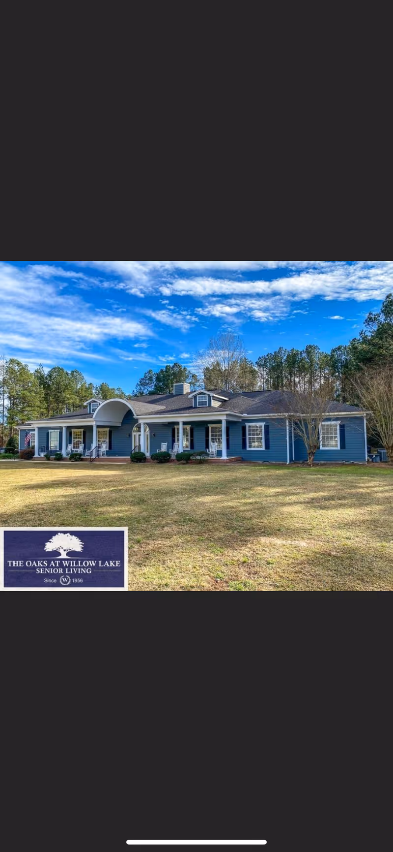 Single-story blue senior living building with a covered front porch and a large grassy lawn under a partly cloudy sky.