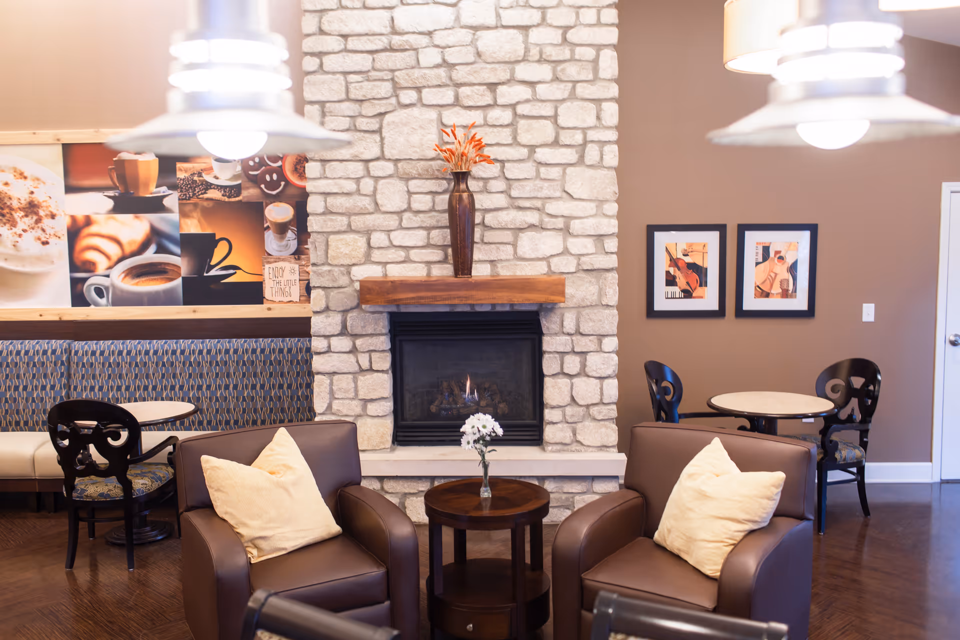Cozy seating area in a senior living facility with two brown leather armchairs each with a beige pillow, a small round wooden table with a vase of white flowers between them, and a stone fireplace with a wooden mantle and a tall vase with orange flowers on top. Behind the seating area are two small round tables with black chairs, a cushioned bench with patterned upholstery, and wall art featuring coffee-themed images and musical instruments.