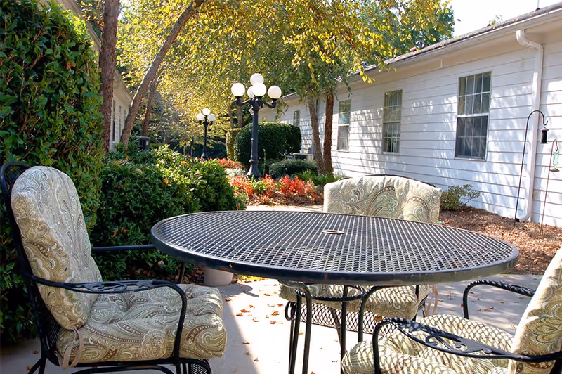 An outdoor seating area featuring a round black metal table surrounded by patterned chairs, with lush greenery and a lamp post in the background.