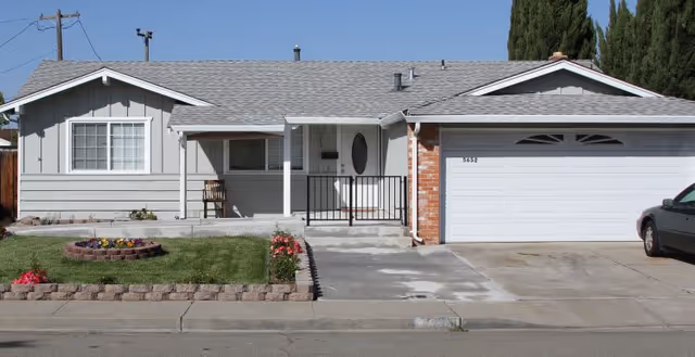 Front exterior of a single-story ranch-style house with a driveway, two-car garage, small front porch, and lawn with flower beds.