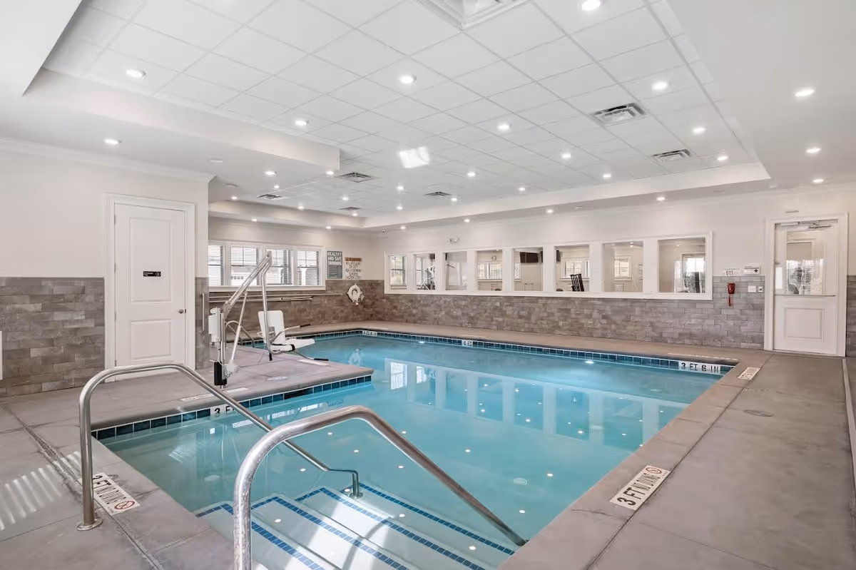 Indoor swimming pool area with clear blue water, metal handrails, a pool lift chair, and a tiled wall with windows looking into an adjacent room. The ceiling has recessed lighting and the floor around the pool is concrete.