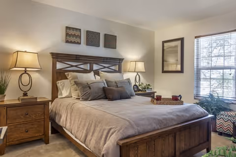 A cozy bedroom with a wooden bed frame and headboard, neatly made with beige and gray bedding and multiple pillows. Two matching wooden nightstands with lamps flank the bed. There are three decorative wall hangings above the headboard and a framed picture on the adjacent wall. A large window with blinds lets in natural light, and there are plants and a patterned ottoman near the window.