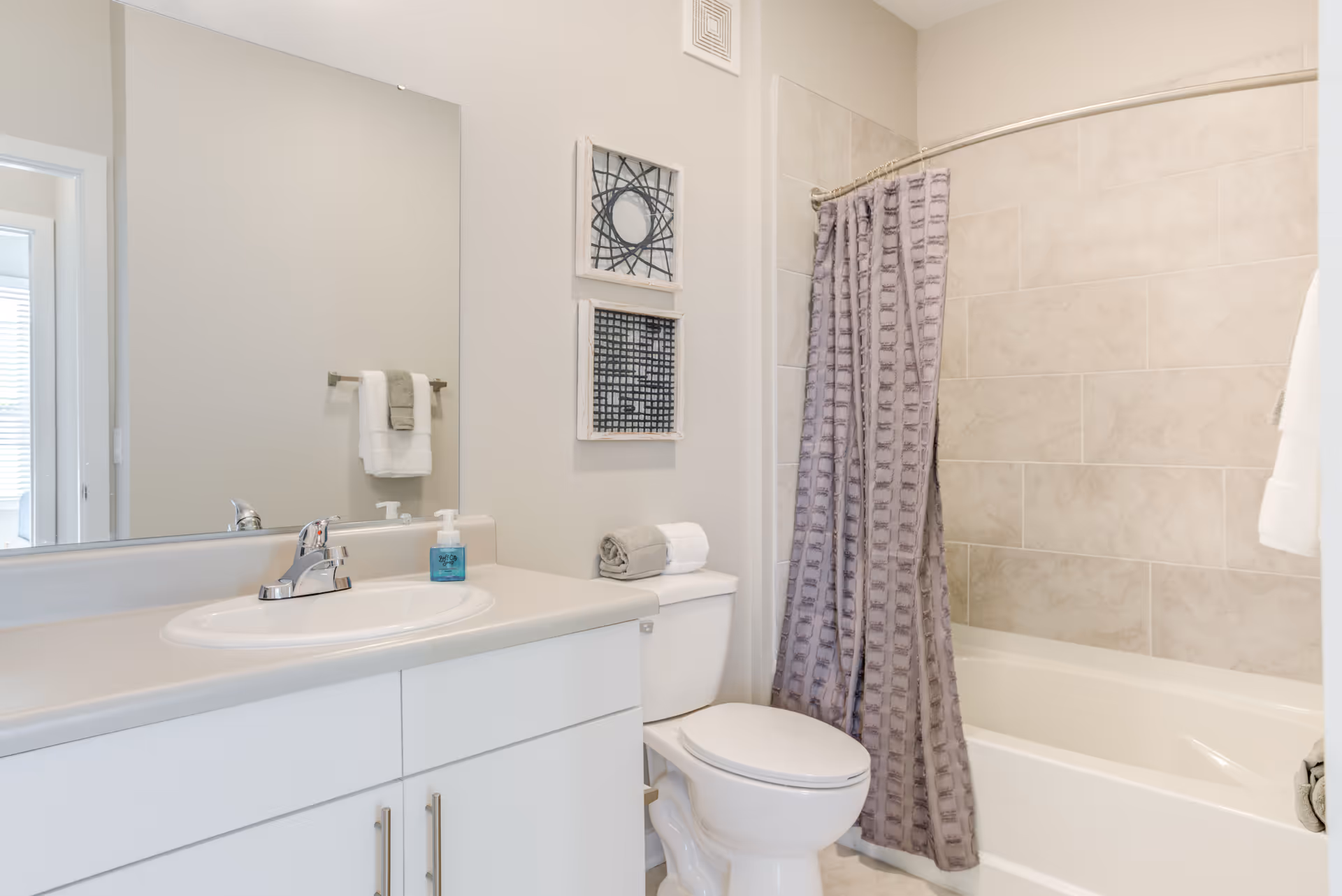 A clean and modern bathroom featuring a white sink with a silver faucet, a large mirror above the sink, a white toilet with neatly folded towels on top, and a bathtub with beige tiled walls and a purple patterned shower curtain.