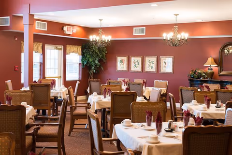 A dining room with multiple tables covered in white tablecloths, each set with cups, plates, and folded burgundy napkins. The room has brown walls adorned with framed botanical prints and a large green plant in the corner. There are chandeliers hanging from the ceiling and a sideboard with a lamp and floral arrangement.