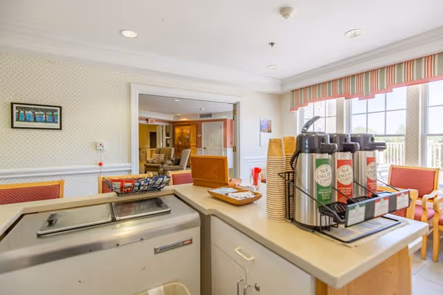 Interior view of a senior living facility's refreshment area with a counter holding several airpots of coffee or tea, disposable cups, and condiments. In the background, there is a seating area with chairs and large windows letting in natural light.