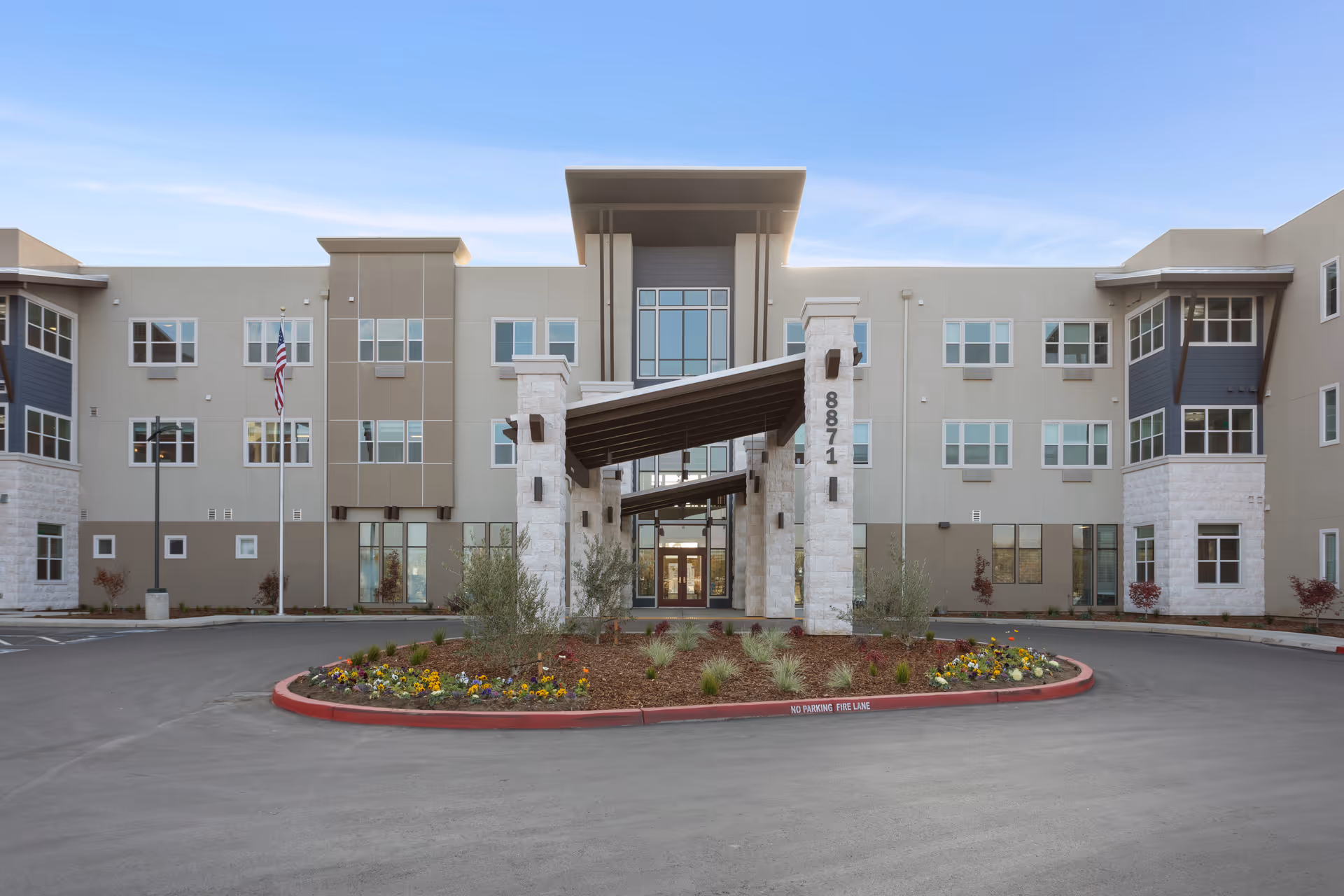 Front exterior view of WellQuest of Elk Grove, a modern three-story building with large windows, a covered entrance supported by stone pillars, and a landscaped roundabout with flowers and shrubs in front.