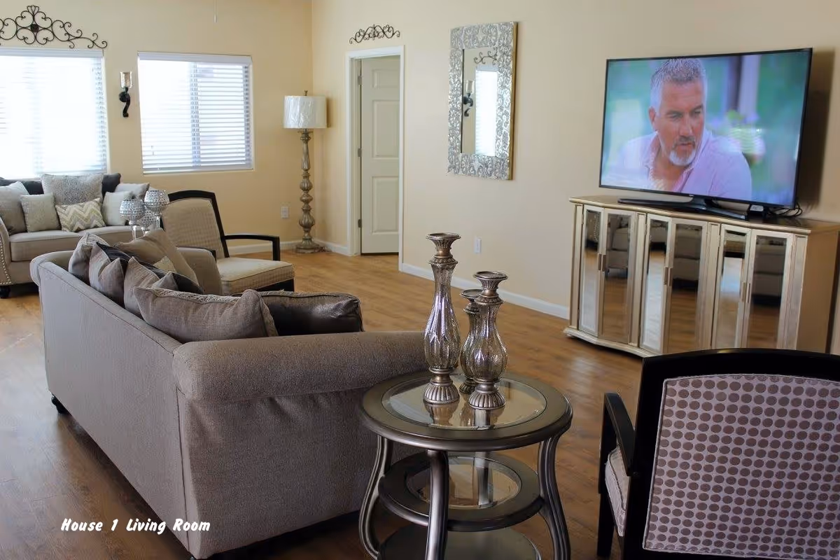 A cozy living room with beige walls and wooden flooring featuring a gray sofa with multiple cushions, a round glass-top side table with decorative vases, a patterned armchair, a floor lamp, a wall mirror, and a flat-screen TV on a mirrored cabinet. Two windows with white blinds allow natural light into the room.