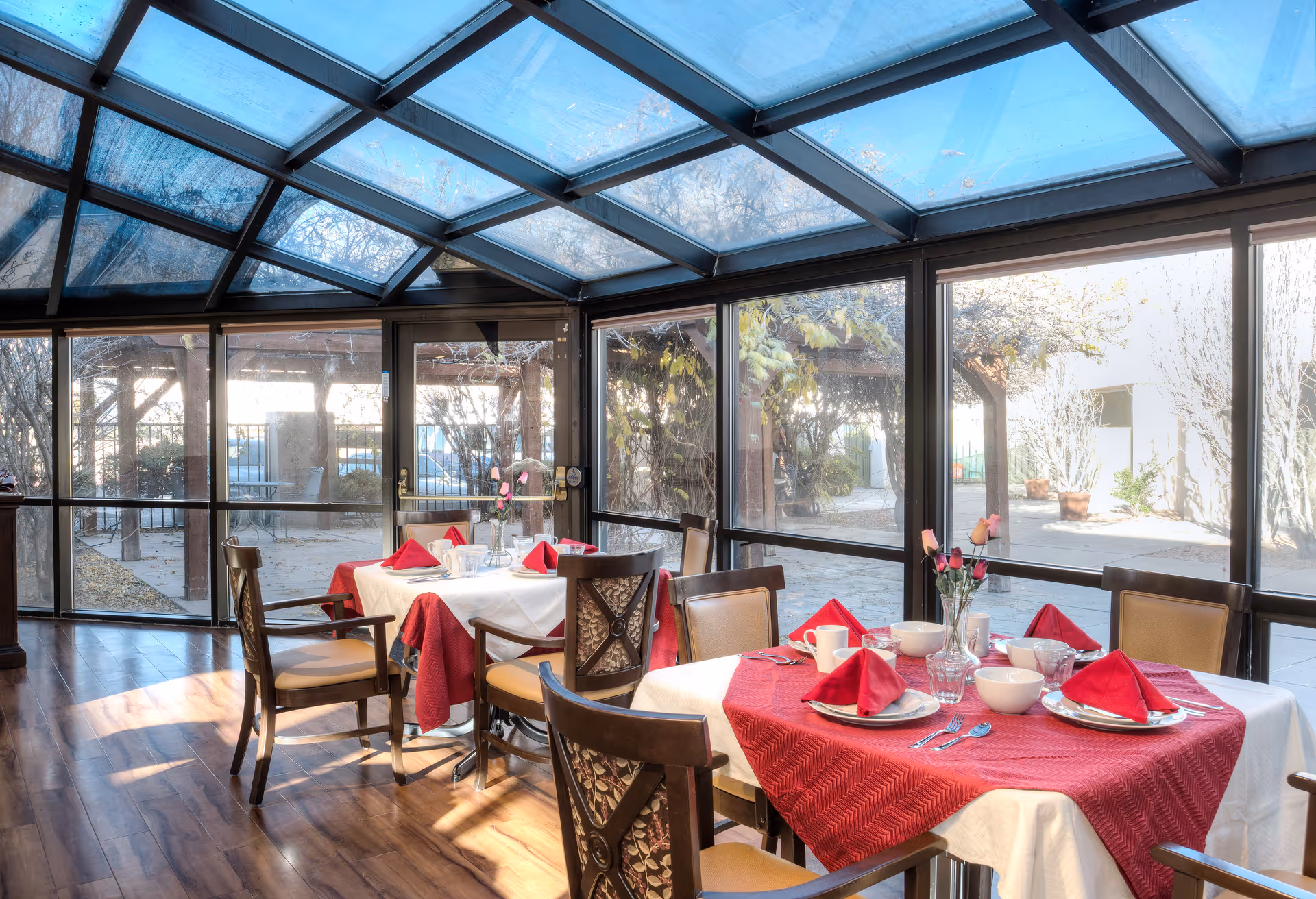 A bright dining area with large glass windows and a glass ceiling, allowing natural light to fill the space. Two tables are set with white tablecloths and red table runners, each adorned with red folded napkins, plates, cups, and a vase with pink roses. Wooden chairs with cushioned seats surround the tables. Outside, there is a patio area with trees and plants.