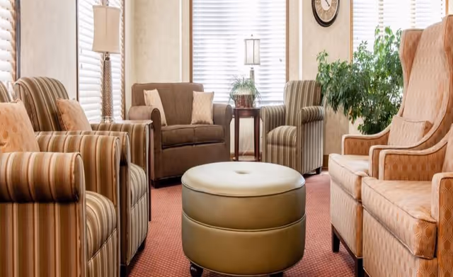 A cozy senior living room with a variety of upholstered armchairs and a loveseat arranged around a round, green ottoman. The room features large windows with white blinds, a side table with a lamp and a plant, a wall clock, and a large leafy green plant in the corner. The carpet is a warm reddish color.