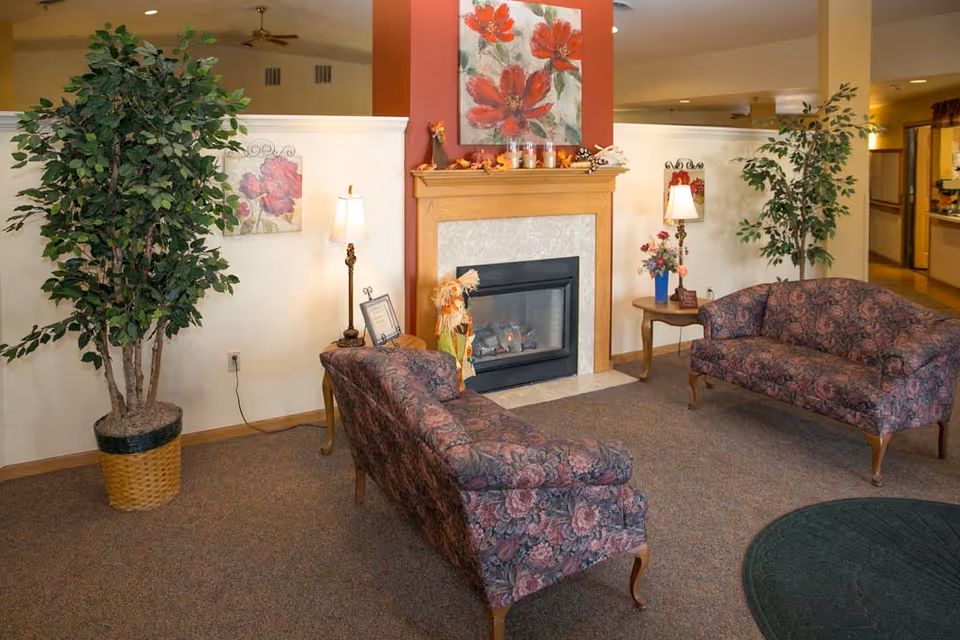 A cozy senior living facility common area with two floral-patterned sofas facing a fireplace. The fireplace has a wooden mantle decorated with candles and autumn-themed items. There are two tall potted plants on either side of the room, two table lamps on small wooden side tables, and floral artwork on the walls. The carpeted floor and warm lighting create a welcoming atmosphere.