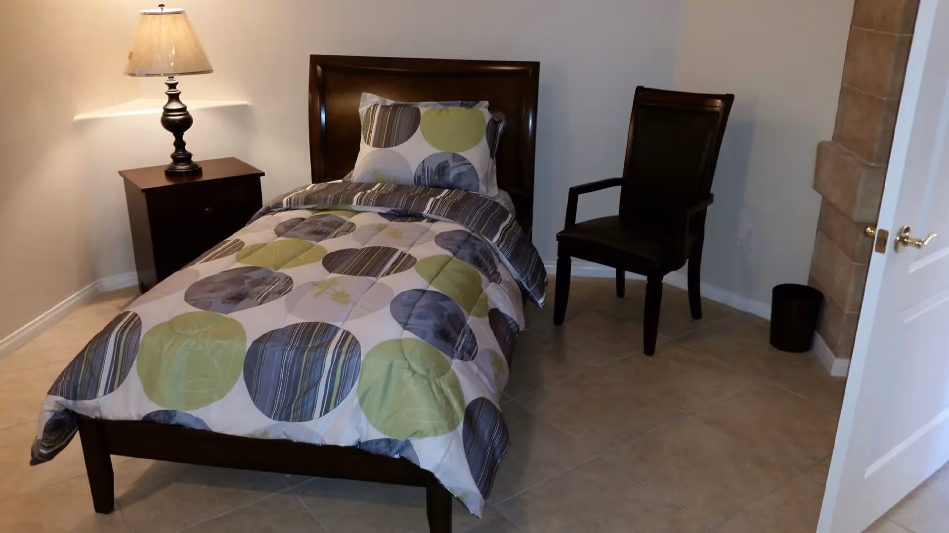 A small bedroom with a single bed featuring a colorful circular patterned comforter and pillow. Next to the bed is a dark wooden nightstand with a lamp on it. There is a dark wooden chair with armrests placed against the wall. The floor is tiled, and a white door is partially open on the right side of the image.