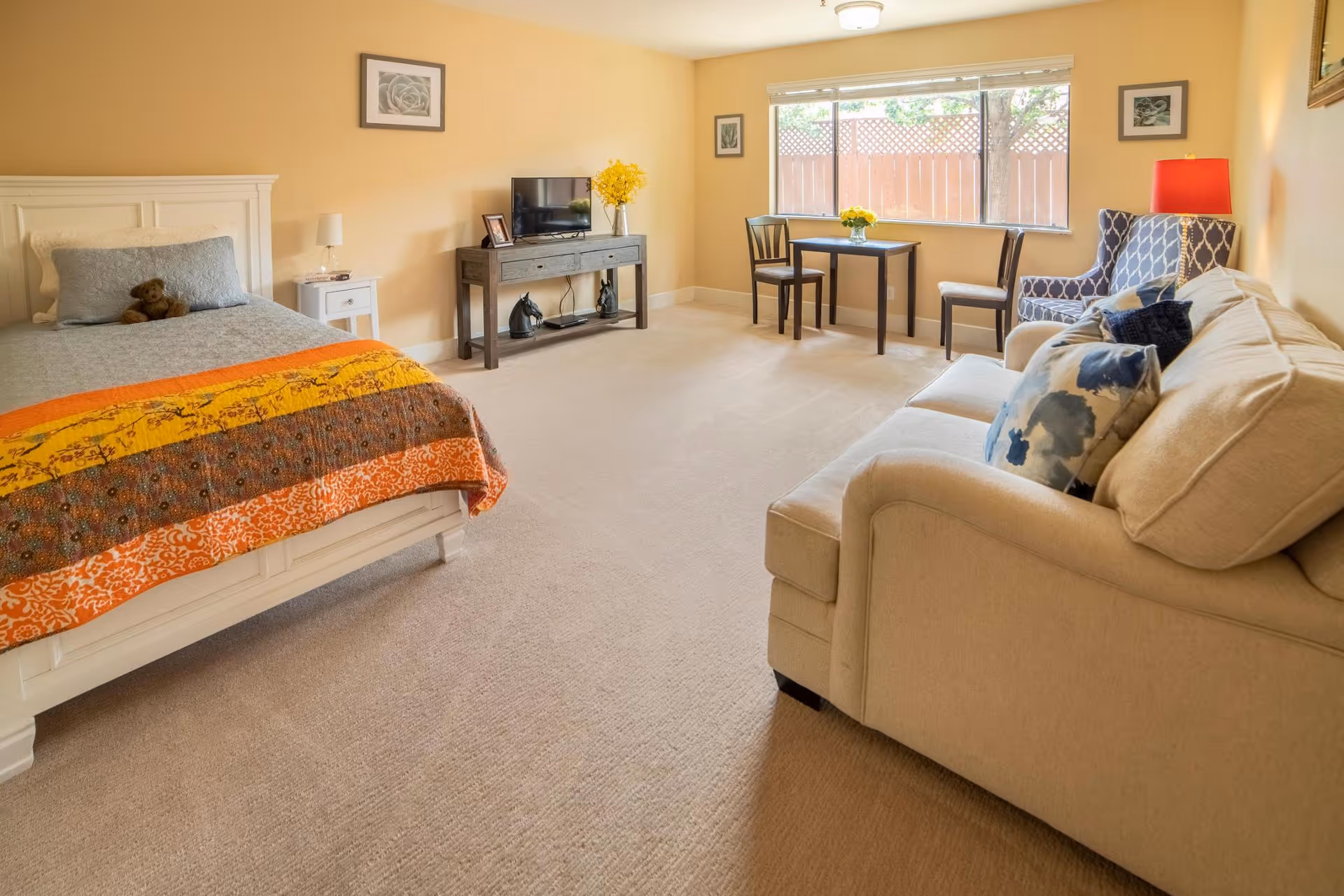 Bright studio room with a bed, sofa, TV on a console, and a small dining table by a large window.