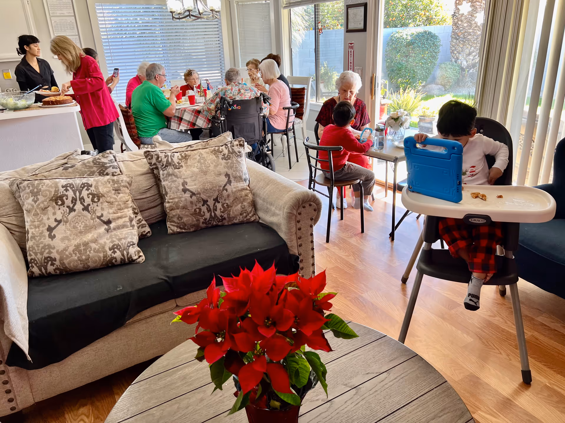 A lively indoor scene at an assisted living home showing elderly residents and children interacting and dining together. In the foreground, there is a beige couch with patterned pillows and a round wooden table with a red poinsettia plant. In the background, several elderly people are seated around a table covered with a checkered tablecloth, eating and talking. Two children are seated at a smaller table near a large window with vertical blinds, one in a high chair using a tablet. The room is bright with natural light coming through the window, and a kitchen counter with two women serving food is visible on the left.
