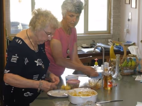 Two elderly women preparing food together in a kitchen. One woman is peeling or cutting a potato while the other is washing or handling something near the sink. Various kitchen items, including a bowl with food, a basket of bananas, and bottles, are visible on the counter.