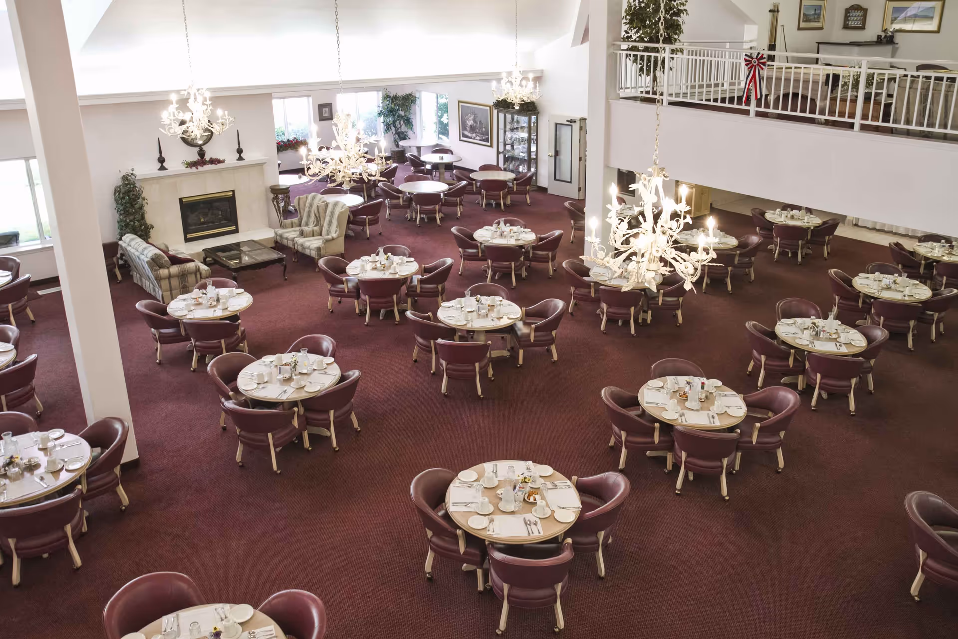 A spacious dining room with multiple round tables set for meals, each surrounded by four maroon chairs. The room features a maroon carpet, chandeliers hanging from the ceiling, a fireplace with a mantel, and a seating area with sofas and armchairs near the windows. There is a second-floor balcony overlooking the dining area.