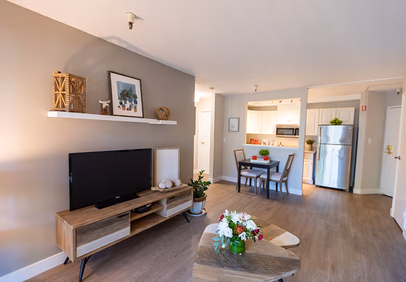 A bright and modern living area in a senior living facility featuring a wooden TV stand with a flat-screen TV, decorative items on a wall-mounted shelf, a small dining table with two chairs, and a kitchen with white cabinets and stainless steel refrigerator in the background. A wooden coffee table with a floral arrangement is in the foreground.