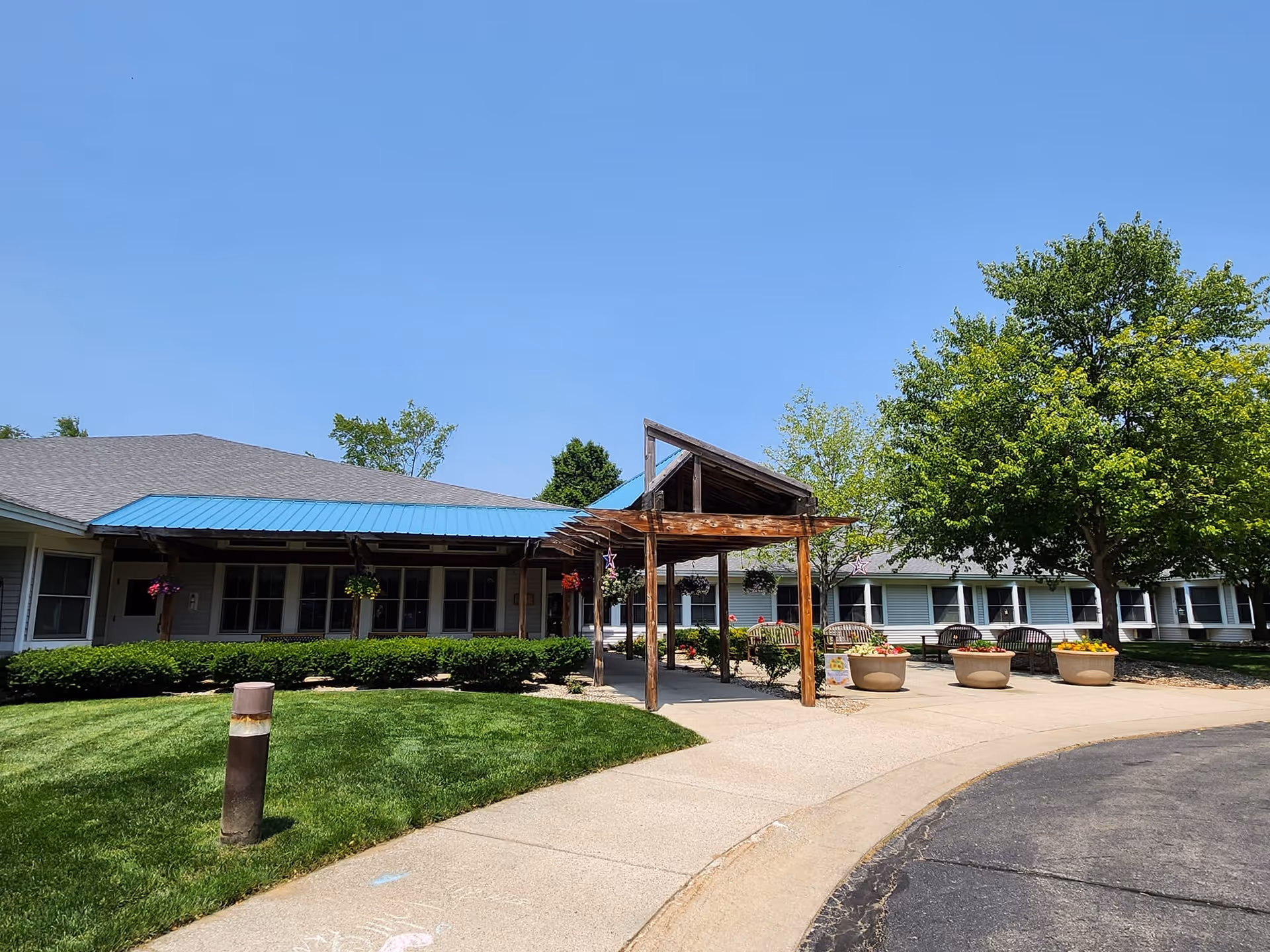 Exterior view of Carveth Village LLC showing a single-story building with a blue metal roof and a wooden pergola over the entrance. There are green bushes, a well-maintained lawn, several large flower pots with colorful flowers, and a large tree providing shade near the entrance. The sky is clear and blue.