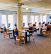 A bright dining room with multiple wooden tables and chairs arranged neatly. Large windows with white curtains allow natural light to fill the space. The room features a carpeted floor and a central column, creating a welcoming atmosphere.