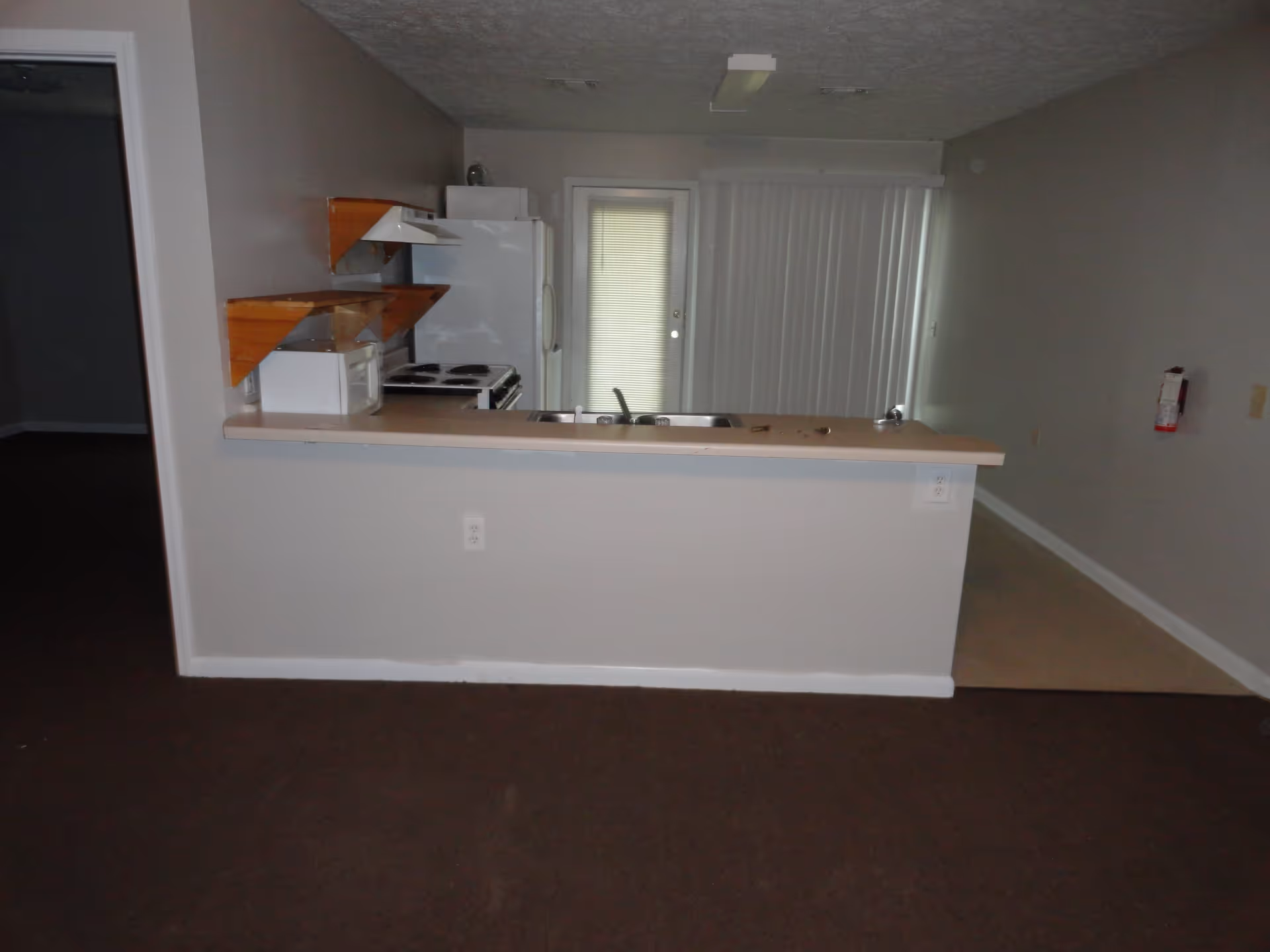 Interior view of a small kitchen area with a countertop and sink in the foreground, white refrigerator and stove against the back wall, wooden shelves mounted on the left wall, and a door with vertical blinds leading outside.