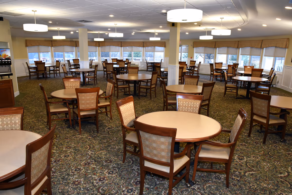 A spacious dining room with multiple round tables surrounded by wooden chairs with patterned upholstery. The room has large windows with beige blinds, carpeted floors with a floral pattern, and ceiling lights providing bright illumination. A beverage dispenser is visible on the left side of the image.