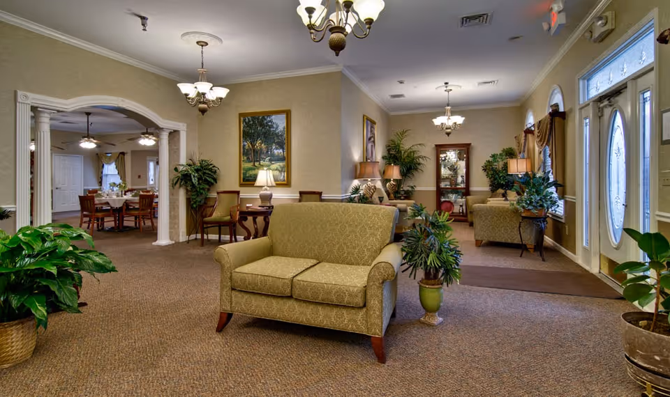 A cozy and well-lit living room area in a senior living facility with beige patterned armchairs, several potted plants, framed paintings on the walls, and multiple table lamps. The room has carpeted floors and elegant chandeliers hanging from the ceiling. In the background, there is a dining area with wooden tables and chairs visible through a large archway.