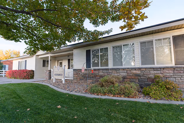Exterior view of Ashley Manor Memory Care, a single-story building with white siding and stone accents. The entrance is visible with a sign displaying the facility's name and phone number. There are green lawns, shrubs, and trees with autumn leaves surrounding the building.