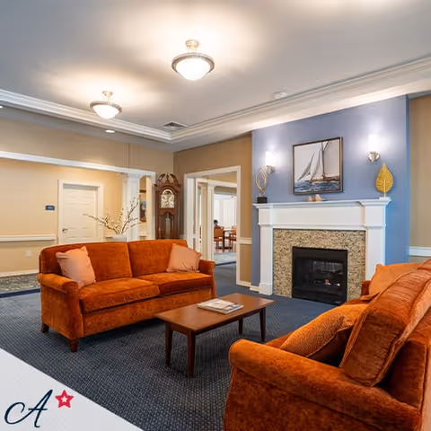 A cozy living room area with two burnt orange sofas facing each other across a wooden coffee table. The room features a fireplace with a stone surround and a mantel decorated with a framed sailboat picture and two decorative items. The walls are painted in beige and blue tones, and the ceiling has recessed lighting fixtures. In the background, there are doorways leading to other rooms, and a grandfather clock is visible near one doorway.