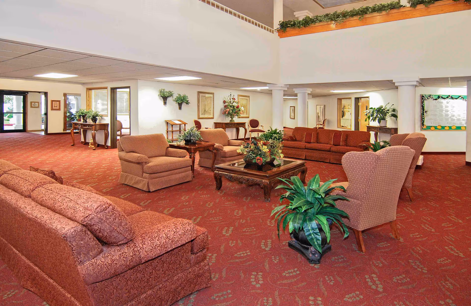 Spacious interior lounge with red carpeting, several upholstered sofas and chairs arranged around coffee tables and potted plants.