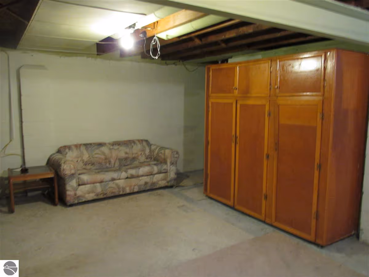 A sparsely furnished basement room with a floral patterned couch, a small wooden side table, and a large wooden wardrobe. The ceiling is partially unfinished with exposed beams and a single light bulb hanging down.