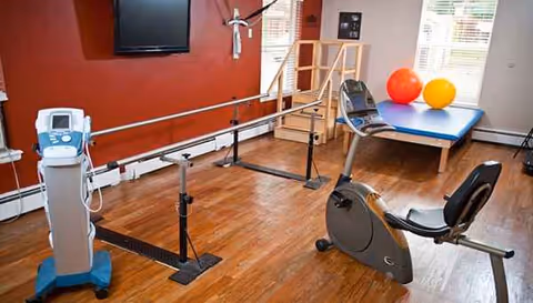 Rehabilitation gym with parallel bars, exercise bike, therapy table, and exercise balls in a senior facility.