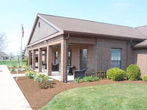 Brick senior living facility with a covered front porch, seating, and manicured landscaping.