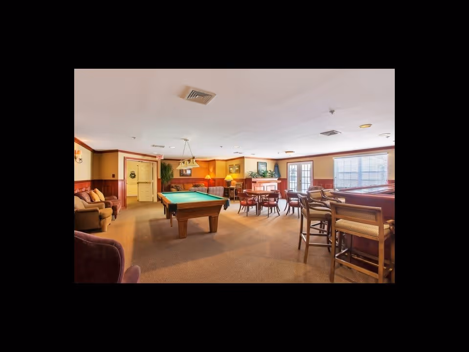 A spacious common room with a pool table in the center, surrounded by comfortable seating including armchairs, a sofa, and bar stools. There are several tables and chairs near a fireplace, with large windows letting in natural light. The room has warm wood paneling and a cozy, inviting atmosphere.