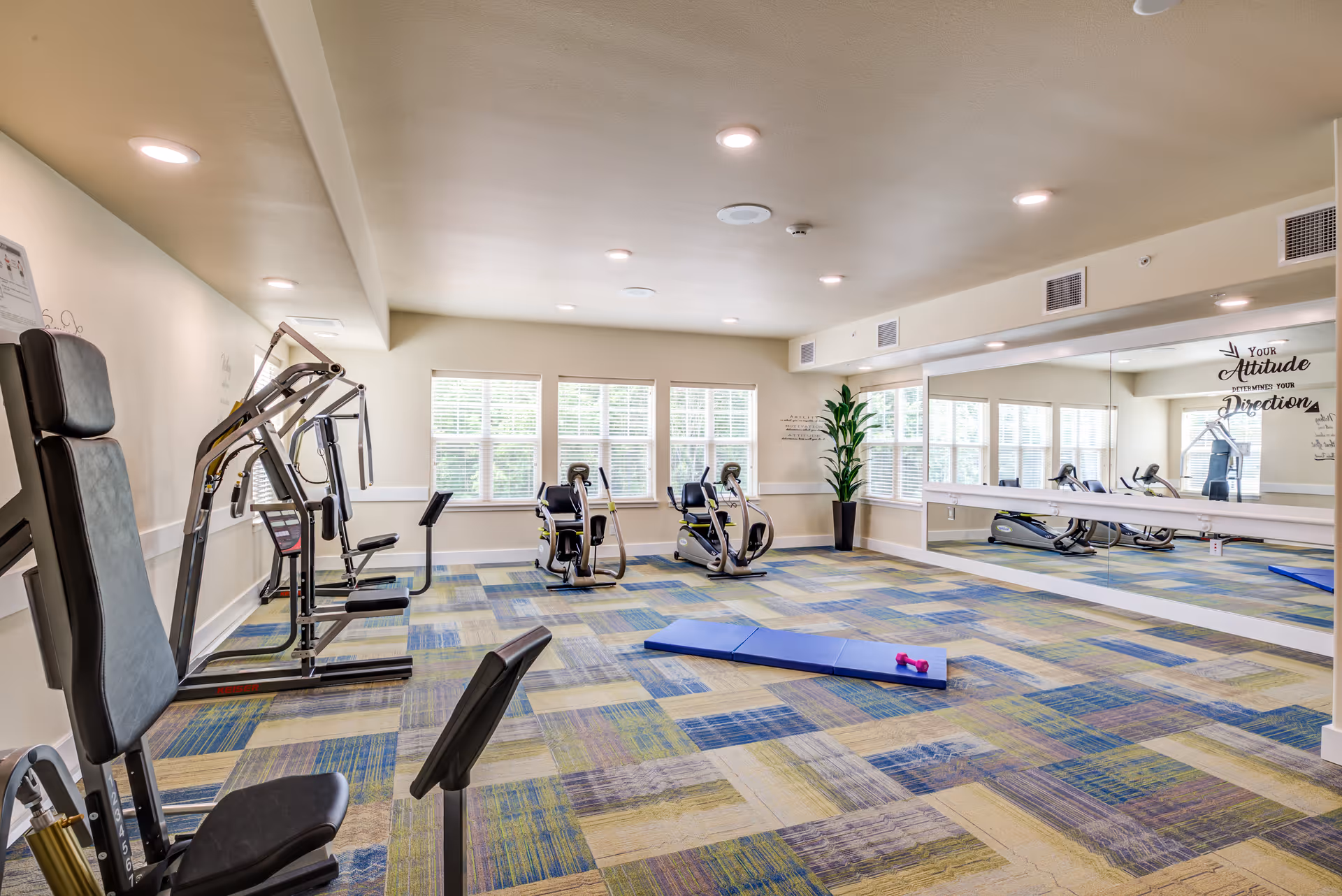 Bright fitness room with exercise machines, a blue exercise mat and a large mirrored wall under recessed lighting.