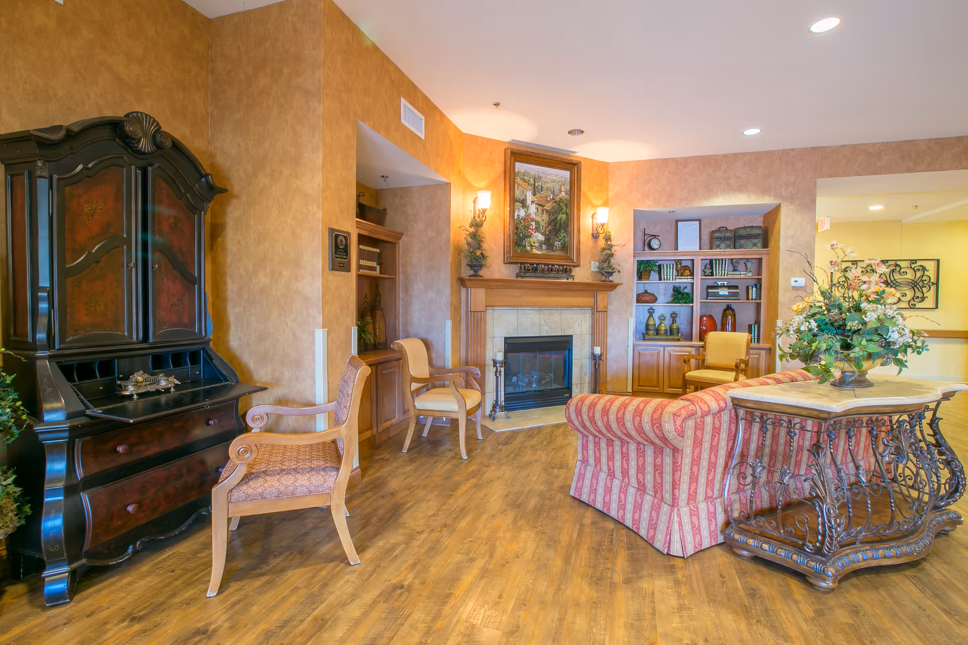 A cozy living room area in a senior living facility with a striped red and beige sofa, a decorative wooden table with a floral arrangement, two wooden chairs with patterned cushions, a fireplace with a painting above it, built-in shelves with books and decorative items, and warm-toned walls and wooden flooring.