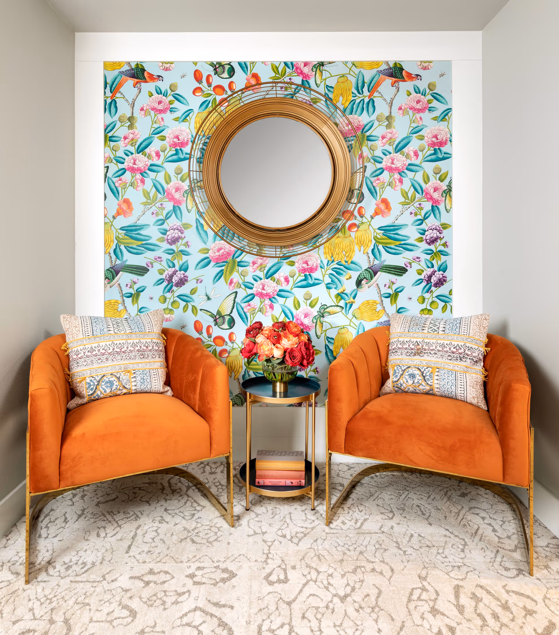 Two orange upholstered armchairs with patterned cushions are placed on a light-colored patterned carpet. Between the chairs is a small round gold and black side table holding a vase of red and orange flowers and some books on the lower shelf. Behind the chairs is a wall with colorful floral and bird wallpaper, featuring a round gold-framed mirror in the center.