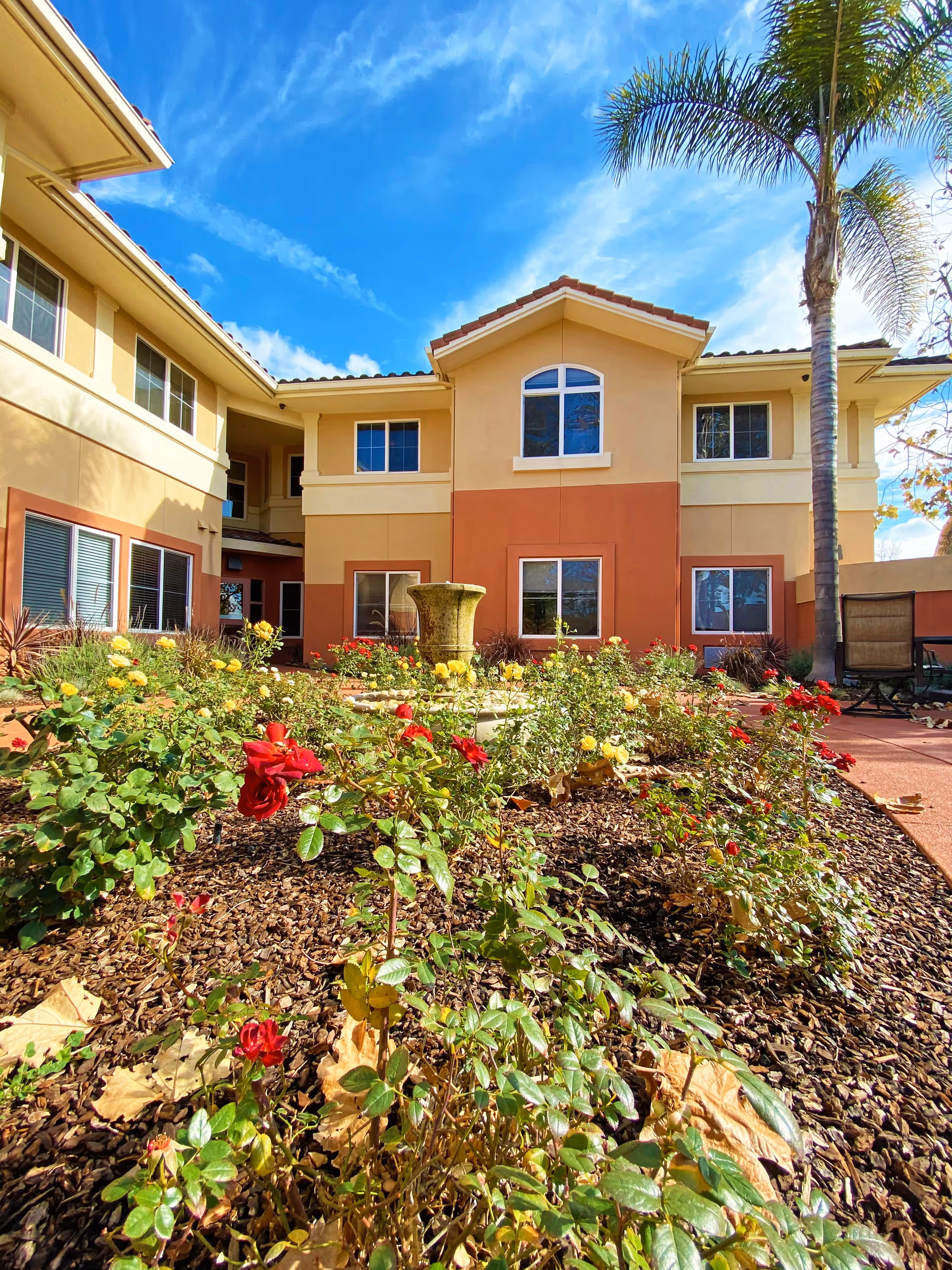 Exterior view of AlmaVia of Camarillo showing a two-story building with beige and terracotta colored walls, multiple windows, a central fountain, and a garden with red and yellow flowers under a bright blue sky with scattered clouds.